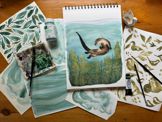 artist, Bevin Marie painting of an otter under the water in a kelp forest, on a table with other paintings and paint and paint brushes