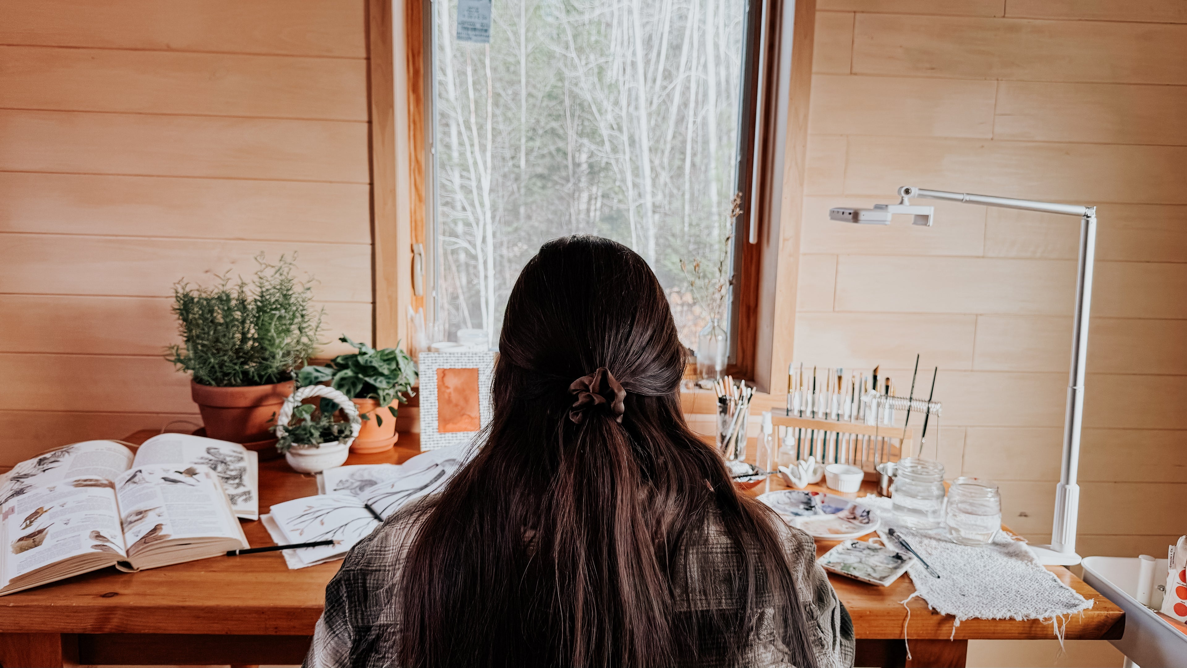 artist, Bevin Marie, sitting at her wooden art desk in her cozy art studio with nature reference books and sketch books open on the table while she is painting water colour.