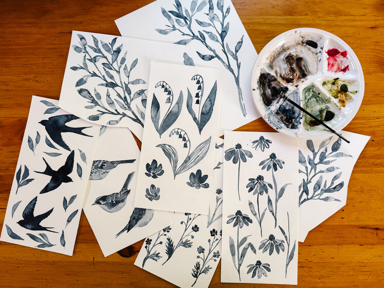Artistic prints of plants and birds on paper with a paint palette and brushes on a wooden surface.